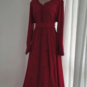 Henna klänning - Elegant vinröd långklänning med vackra broderier och pärlor. Klänningen har långa ärmar och ett matchande bälte i midjan. Perfekt för festliga tillfällen.