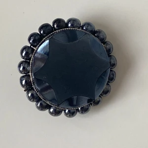 Svart brosch med pärlor - Elegant rund brosch i svart med en central stjärnformad design. Omgiven av glänsande svarta pärlor som ger en lyxig känsla. Perfekt som en stilfull accessoar för att lyfta din outfit.