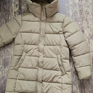 Beige dunjacka med hög krage - Säljer en stilren beige dunjacka med hög krage och knappar. Jackan har en quiltad design och långa ärmar, perfekt för kyliga dagar. Den har praktiska fickor och en dragkedja framtill.