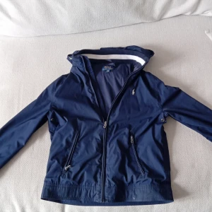 Mörkblå vindjacka från Polo Ralph Lauren - Säljer en stilren mörkblå vindjacka från Polo Ralph Lauren. Jackan har en dragkedja framtill och en broderad logotyp på bröstet. Perfekt för blåsiga dagar med sin lätta och bekväma design.