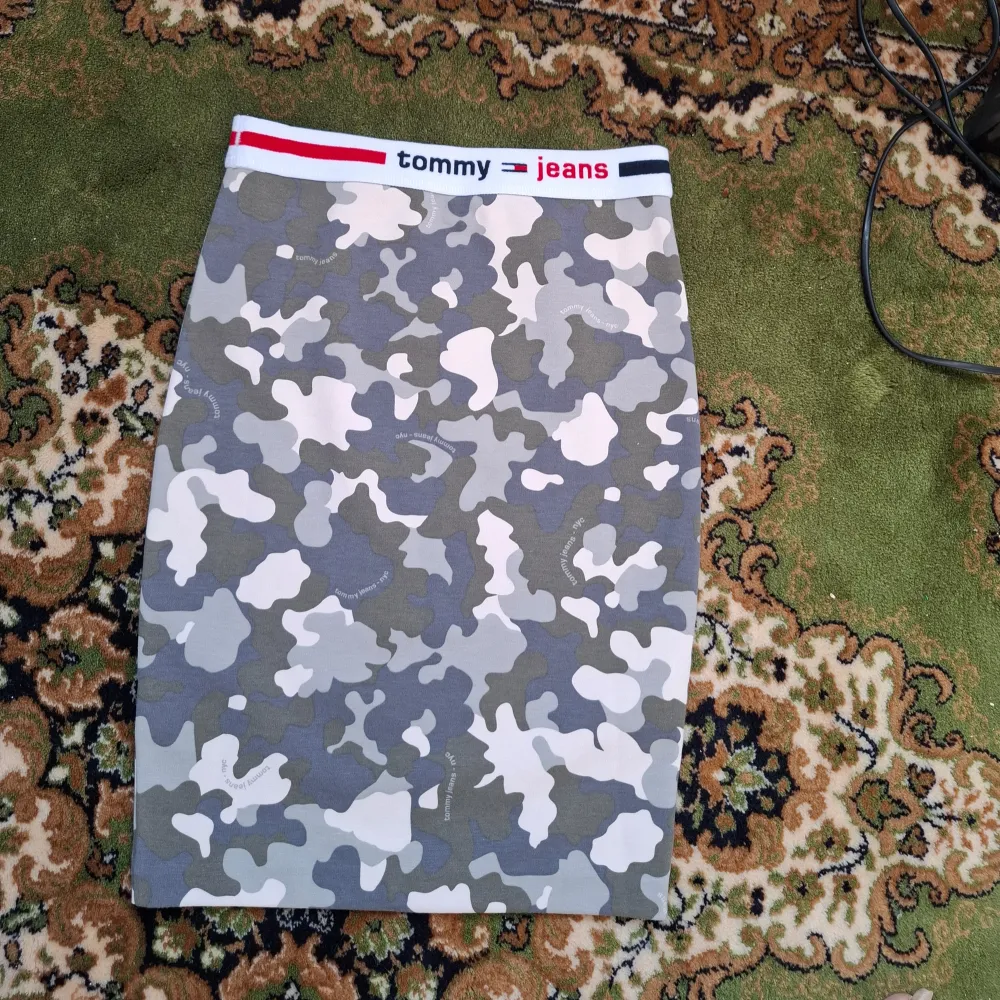 Snygg camo-mönstrad kjol från Tommy Jeans i storlek XS. Kjolen har en elastisk midja med märkets logga i rött och blått. Perfekt för en cool och avslappnad stil.. Hameet.