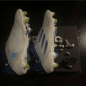 Vita och blå fotbollsskor från Adidas - Säljer ett par vita och blå fotbollsskor från Adidas med snörning och utbytbara dobbar. Skorna har en modern design med Adidas-loggan på sidan och är perfekta för fotbollsplanen.