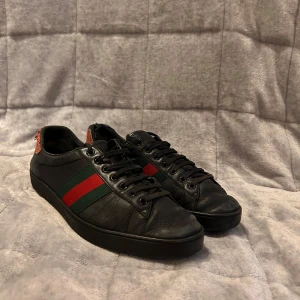 Svarta sneakers med röd och grön rand - Snygga svarta sneakers från Gucci med ikoniska röd och grön rand på sidan. Skorna har en rund tå och snörning framtill. Hälpartiet är i en kontrasterande röd färg med textur. Perfekta för en stilren look. Modell: Gucci ace. Äkthetsbevis: se bild 4-5. Storlek: 42,5. Skriv privat vid frågor, prisförslag eller byten 🍾