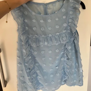 Ljusblå blus med volanger - Söt ljusblå blus med volanger och blommigt mönster. Blusen är ärmlös och har en rund halsringning. Perfekt för en somrig look!