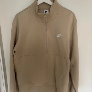 Beige halfzip sweatshirt från Nike - Säljer en beige långärmad sweatshirt från Nike med halv dragkedja och broderad vit logga på bröstet. Tröjan har hög krage och är i mjukt material, perfekt för en avslappnad stil.
