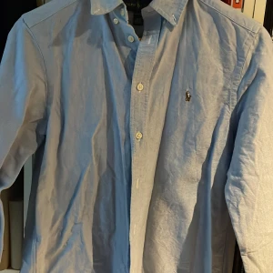 Ljusblå skjorta från Ralph Lauren - Klassisk ljusblå skjorta från Ralph Lauren med broderad logga på bröstet. Skjortan har lång ärm, knappar framtill och en tidlös design som passar till många tillfällen. Tillverkad i bomull för en bekväm känsla. Står 14-16 i strl men passar bäst XS