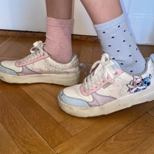Sneakers med Stitch-motiv och pastellfärger - Säljer ett par sneakers i vitt med ljusrosa och ljusblå detaljer samt ett tryck av Stitch och Angel på sidan. Skorna har snörning och en platt sula. Perfekta för dig som gillar Disney och vill ha något unikt på fötterna!