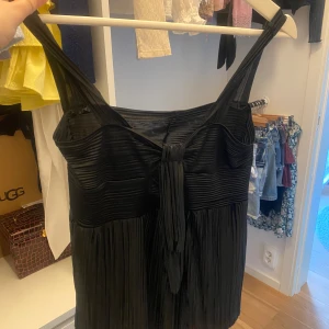 Svart blustopp med knytdetalj - Svart topp med plisserad tyg och breda axelband. Överdelen har horisontella veck och en snygg knytdetalj framtill. Perfekt för dig som gillar en stilren look med det lilla extra. Har en defekt på tyget vid knytet därav priset.