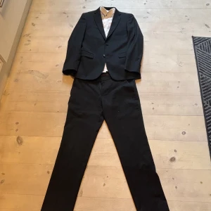 Svart kostym med vit skjorta Lindberg  - Säljer en svart kostym med vit skjorta från Lindberg. Vanlig passform (slim) , komplett med matchande kavaj och byxor samt en vit skjorta. Perfekt för bal eller fest. Kavajen har knappar framtill och skjortan är långärmad med krage. Totalt kostar byxor, kavaj och skjorta runt 3000kr (skjorta 1000kr, kavaj och kostymbyxor 2000kr) väldigt bra skick och knappt använda 9,5/10. Pris för hela kostymen 999. Men de går att köpa sakerna för sig om de skulle önskad. Skriv för funderingar! Storlek s/m