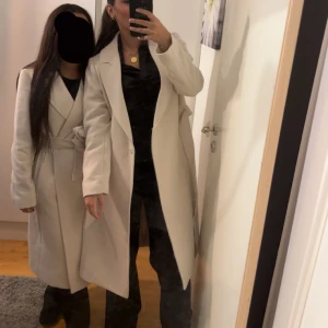 Beige kappa med bälte - Säljer en stilren beige kappa med bälte i midjan. Lång modell med klassisk krage och långa ärmar. Perfekt för dig som vill ha en tidlös och elegant look. Använd en gång och köpt för 799