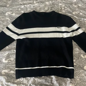 Svart och vit stickad tröja från Lacoste - Säljer en svart stickad tröja från Lacoste med vita horisontella ränder över bröstet och ärmarna. Tröjan har rund halsringning och den klassiska Lacoste-loggan på bröstet. Perfekt för en stilren och sportig look.