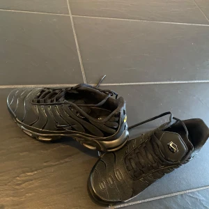 Svarta Nike Air Max Plus TN sneakers - Säljer ett par helsvarta Nike Air Max Plus TN sneakers med klassisk Tn-detalj på hälen. Skorna har snörning, rund tå och en synlig luftdämpad sula. Perfekta för dig som gillar sportig och stilren look.