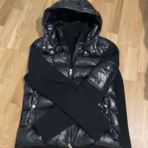 Svart pufferjacka med huva från Moncler - Säljer en svart glansig pufferjacka från Moncler med huva och dragkedja framtill. Jackan har långa ärmar i stickat material och Moncler-logga på ena ärmen. Perfekt för kyliga dagar.