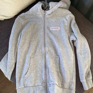 Grå hoodie från Peak Performance - Grå hoodie med dragkedja från Peak Performance. Tröjan har huva, två fickor framtill och en liten logotyp på bröstet. Perfekt för en avslappnad stil och passar året runt.