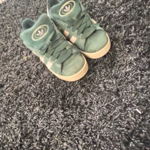 Äkta Adidas Campus sneakers i grön mocka - Säljer ett par Adidas Campus sneakers i grön mocka med vita ränder och beige sula. Skorna har klassisk snörning och Adidas-logga på plösen. Perfekta för dig som gillar retrostil och vill ha något unikt i garderoben.