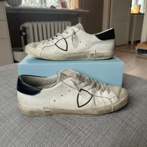 Philippe Model PRSX - Säljer ett par vita Philippe Model sneakers med svarta detaljer på hälen och gråa partier. Skorna har snörning och en klassisk låg profil. Perfekta för dig som gillar stilrena och trendiga sneakers. Skorna är i bra skick dock några små slitage/skador men som eventuellt går att fixa. Köpta hösten 2024 men används inte längre. Box och dustbag medföljer. Hör av er vid funderingar! Pris kan diskuteras