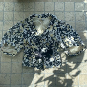 Blommig kortärmad kavaj med rosett - Säljer en unik blommig kavaj från Jesire med svart och gul mönster, korta ärmar och stor krage. Jackan har knappar framtill och en dekorativ svart rosett i midjan. Skulle använt den själv om jag visste hur man ska styla den. Det står att den är dry clean men jag körde den i vanlig tvätt och den kom ut helt oskadd.