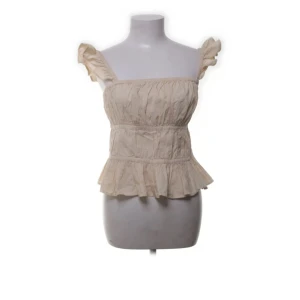 Beige volangtopp med resår - Söt beige topp med volangdetaljer och resår i midjan. Toppen har breda axelband med volang och en lätt rynkad struktur. Perfekt för varma dagar och ger en romantisk känsla till outfiten.