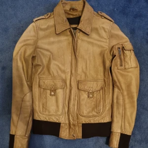 Beige skinnjacka - Säljer en beige skinnjacka med avtagbar pälskrage. Jackan har dragkedja, fickor med lock och knappar samt en ärmficka med dragkedja. Jackan har en svag rosa fläck som syns på höger sida (sista bilden)