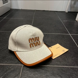 Beige keps från Miu Miu - Snygg beige keps från Miu Miu med småprickigt mönster och bruna detaljer längs skärmen och justerbart spänne bak. Broderad Miu Miu-logga framtill i brunt och vitt. Perfekt accessoar för en trendig look.