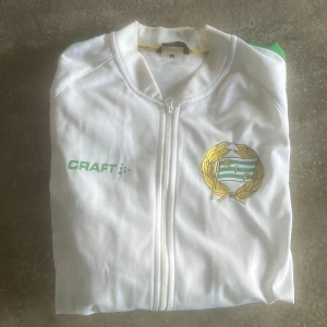 Vit Hammarby track jacket från Craft - Säljer en vit track jacket från Craft med Hammarby-emblem broderat på bröstet och gröna detaljer längs ärmarna. Dragkedja framtill och stor grön 'HAMMARBY'-text på ryggen. Perfekt för dig som älskar fotboll och vill visa ditt stöd för laget.💚🤍