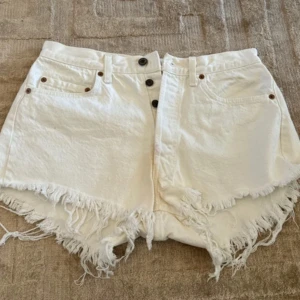 Vita jeansshorts med fransar från Levi's - Säljer ett par vita jeansshorts från Levi's med råa fransade kanter och klassisk femficksdesign.Strl. Ca M se bild med mtt. Shortsen har knappar framtill och bälteshällor. Perfekta för sommaren och riktigt snygga till sneakers eller sandaler. Finns fläck framme, se bild. Tror går bort med lite blekmedel