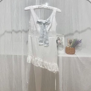 Vit ärmlös klänning med spets och rosett - Supersöt vit klänning med asymmetrisk volangkant och spetsdetaljer. Baksidan har en stor ljusblå rosett som ger en gullig touch. Passar även XXS. Om du är över 150cm blir det en topp istället.