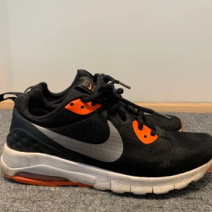 Svarta Nike sneakers med orange detaljer - Säljer ett par svarta Nike sneakers med orange detaljer och vit sula. Skorna har en stor silverfärgad swoosh på sidan och svarta skosnören. Perfekta för träning eller vardag, med bekväm passform och sportig look.