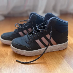 Mörkblå Adidas sneakers med rosa ränder - Säljer ett par mörkblå Adidas sneakers med klassiska rosa ränder på sidorna. Skorna har högt skaft, svart-vita snören och vit sula. Insidan är fodrad med mjukt material för extra komfort. Perfekta för dig som gillar sportig stil.