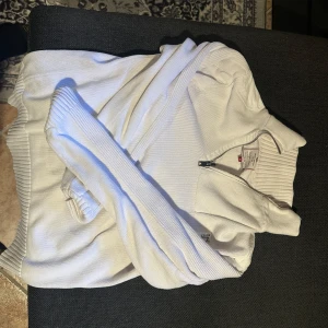 Vit ziptröja från Jack & Jones - Säljer en vit långärmad ziptröja från Jack & Jones med tryck på ryggen och ribbade muddar. Tröjan har hög krage och dragkedja framtill. Perfekt för en avslappnad stil.