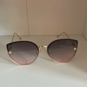 Fendi solglasögon med guldiga detaljer - Snygga solglasögon från Fendi med rundad cat-eye form. Glasen är mörktonade i grått och bågarna är i guldmetall med Fendi-logga på sidorna. Tunn och elegant design som ger en lyxig känsla.