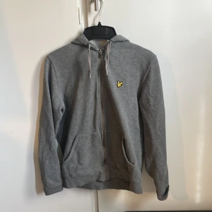 Grå hoodie från Lyle & Scott - Säljer en klassisk grå hoodie från Lyle & Scott med dragkedja och huva. Tröjan har två fickor framtill och den ikoniska gula loggan på bröstet. Perfekt för en avslappnad stil. Orginal pris är 799 mitt pris är 400, priser går att diskutera !