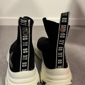 Svarta sock sneakers från Steve Madden - Säljer ett par svarta sock sneakers från Steve Madden med vit sula och tydlig logga på sidan. Skorna har en strumpliknande passform och breda svarta remmar med vit text baktill. Perfekta för en trendig och sportig look. Använt ca. 2-3 gånger💕