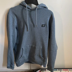 Blå hoodie från Les Deux - Säljer en stilren blå hoodie från Les Deux med broderad logga på bröstet. Tröjan har huva med dragsko och en stor magficka framtill. Perfekt för en avslappnad och snygg look.Orginal pris är 1399kr 