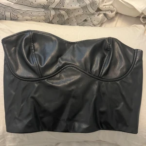 Svart skinn tubtopp - Säljer en svart tubtopp i skinnimitation. Toppen är axelbandslös. Storlek S från Ginatricot. 