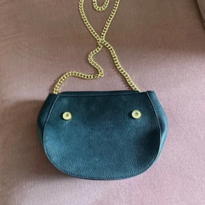 Svart axelväska med guldkedja  - Stilren svart axelväska i skinn med guldfärgad kedja som axelrem. Väskan har rundad form, lock och magnetknappar samt innerfack med dragkedja. Perfekt för att bära det viktigaste med stil.