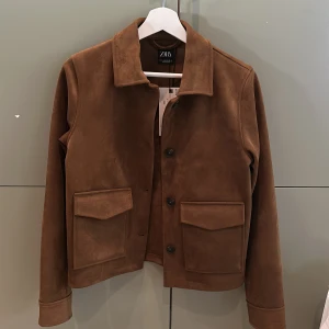 Brun mockajacka från Zara - Säljer en brun mockajacka från Zara med klassisk krage och stora fickor framtill. Jackan har knäppning med svarta knappar och raka ärmar. Perfekt för dig som gillar stilren och tidlös design. Helt ny med prislapp.