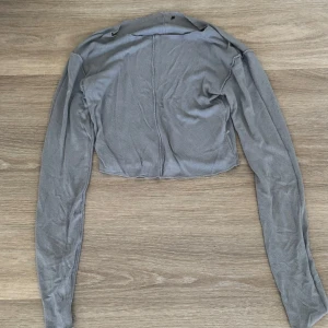 Grå croppad bolerotopp - Säljer en grå croppad bolerotopp med lång ärm. Tunn och mjuk känsla, perfekt att slänga över ett linne eller klänning. Enkel och stilren design utan synliga detaljer framtill.