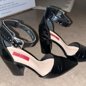 Svarta klackskor från London Rebel - Säljer ett par svarta klackskor från London Rebel med krokodilpräglat mönster. Skorna har spetsig tå, bred klack och justerbart ankelband med spänne. Har några skråmor vid tån och sulan är lite smutsig.  Kan skicka fler bilder :) priset går att förhandla