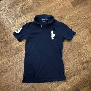 Mörkblå pikétröja från Polo Ralph Lauren - Säljer en mörkblå pikétröja från Polo Ralph Lauren med vit broderad logga på bröstet och siffra på ärmen. Slim fit-modell med klassisk krage och två knappar framtill. Perfekt för en stilren och sportig look. Säljer på grund av att det inte är min stil. Fick av brorsan. Det är min storlek så det är bara att skriva om du funderar över något😁Pris kan alltid diskuteras