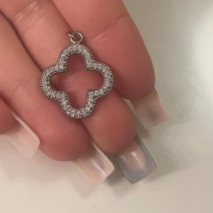 Silverfärgat fyrklöverformat hänge med stenar - Säljer ett snyggt hänge i form av ett fyrklöver, dekorerat med glittrande stenar runt hela kanten. Perfekt att använda som berlock till halsband eller armband. Ger en elegant touch till din stil.
