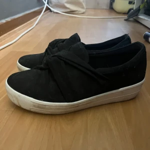 Svarta slip-on sneakers med knytdetalj - Svarta slip-on sneakers med en omlott/knyt design. Skorna är i textilmaterial. Säljer då de är för små och inte kommer till användning. Pris går att diskutera💕
