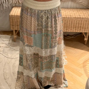 Somrig långkjol  - Säljer denna långkjol  i pastelliga färger. Kjolen har bred smock i midjan och inslag av broderade spetsdetaljer. Använd typ 1 gång så i fint skick! 