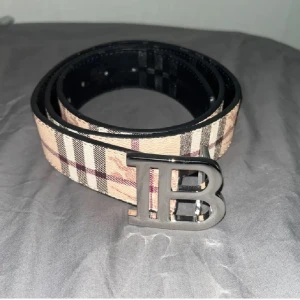 Rutig bälte med metallspänne från Burberry - Snyggt bälte med klassiskt rutigt mönster i beige, svart, vitt och rött. Bältet har ett stort metallspänne med bokstäverna 'B'. Perfekt accessoar för att lyfta din outfit.