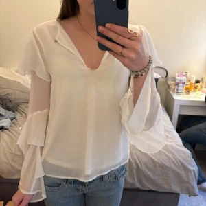 Vit blus med volangärmar - Säljer en luftig och elegant vit blus med v-ringning och långa ärmar med volangdetaljer. Blusen är tillverkad i ett tunt, lätt material som ger ett flowy intryck. Perfekt för dig som gillar romantiska och feminina plagg.