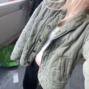 Olivgrön quiltad jacka från Zara - Säljer en olivgrön quiltad jacka från Zara med rund halsringning och knäppning framtill. Jackan har två fickor och ett vågigt quiltat mönster. Perfekt för lager-på-lager och en snygg vardagslook.