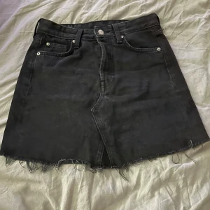 Svart jeanskjol från H&M - Säljer en svart jeanskjol från H&M i storlek 32. Kjolen har rå kant nertill, klassiska fem fickor och knäppning med  knapp framtill. Perfekt för dig som gillar en enkel men snygg stil.