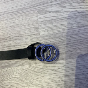 Svart barn bälte med dubbla ringar - Svart barn bälte med två silverfärgade ringar. Bältet är 55cm långt och använt ett tag, det är bra skick förutom en liten skada på slutet av bäletet men det syns inte på utsidan. 