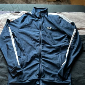 Blå träningsjacka från Under Armour - Snygg blå träningsjacka från Under Armour med vita ränder längs ärmarna och dragkedja framtill. Jackan har hög krage och ribbade muddar vid ärmslut och nederkant. Perfekt för sportiga tillfällen.