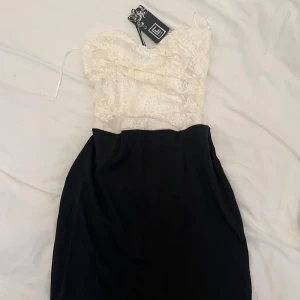 Vit och svart klänning från club L - Säljer en elegant kort klänning från club L med vit spets upptill och svart kjoldel. Axelbandslös design och figurnära passform. Perfekt för speciella tillfällen.